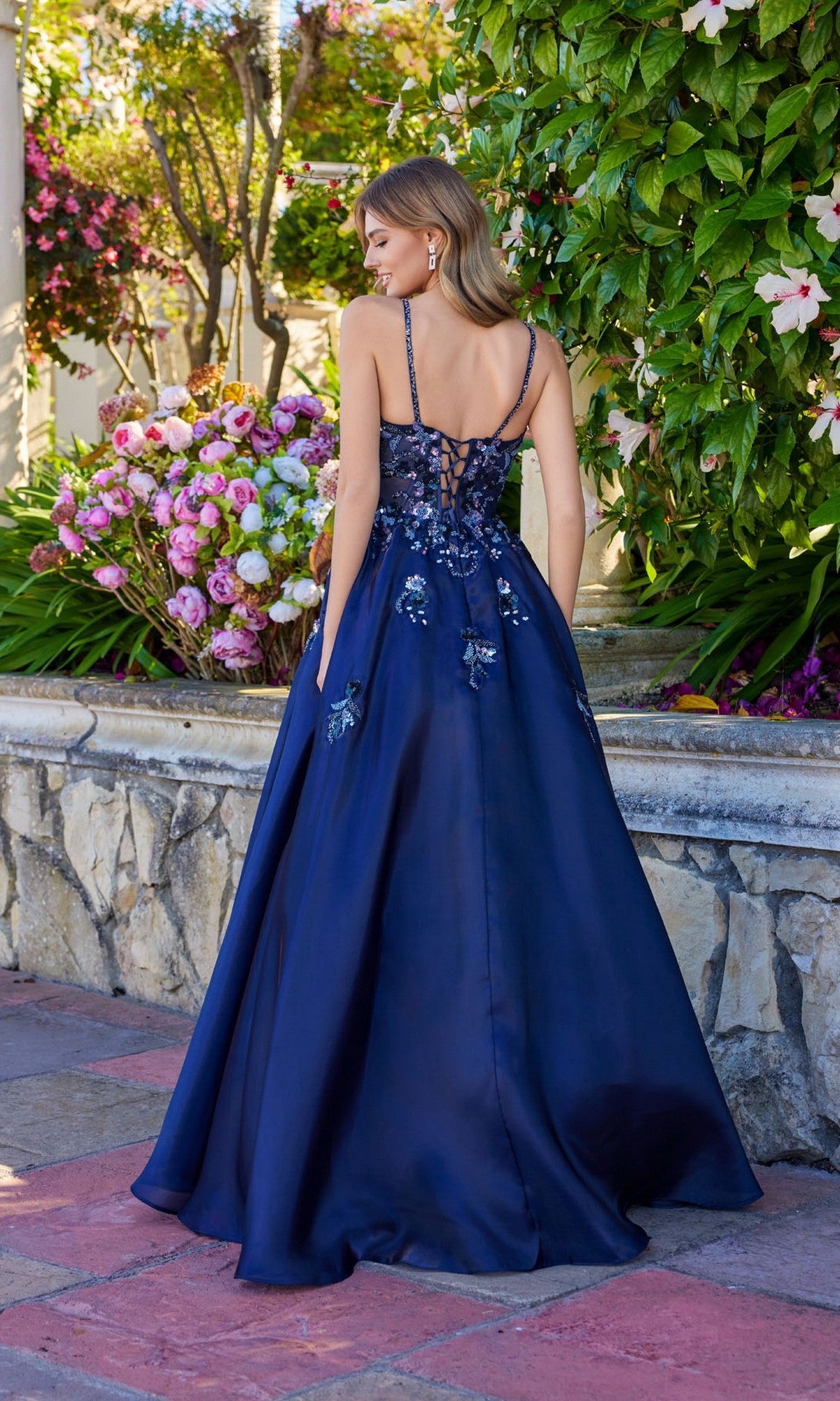 A woman stands outdoors near a stone wall and flowers, wearing the Juliet JT2617U long prom dress—a dark blue A-line gown with floral sequin embroidery and a lace-up back.